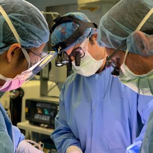 Three male surgeons wearing blue scrubs, surgical maskes and surgical loupes perform procedure in hospital operating room