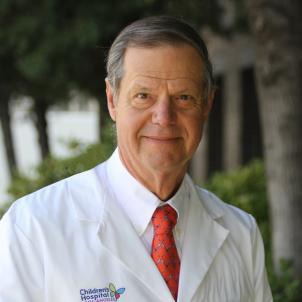 Headshot of a man with light skin tone and grey hair wearing a white dress shirt and red tie beneath a white doctor's lab coat against a blurred outdoor background