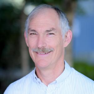 Headshot of a smiling man with light skin tone and short grey hair wearing an open-collared, light colored, striped dress shirt against a blurred outdoor background