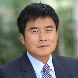 Headshot of a man with medium skin tone and short dark hair wearing light blue dress shirt and purple striped tie under a dark suit jacket against a blurred outdoor background