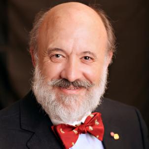 Headshot of a smiling man with light skin tone, a grey beard and receding hairline wearing a light dress shirt and red bow tie under a dark suit jacket against a dark indoor background