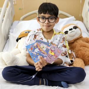 Boy with medium skin tone and dark hair wearing glasses, hospital gown and sweatpants holds three colorful Valentine's Day cards as he sits in his hospital bed next to a stuffed sloth toy