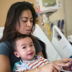 A medium skin-toned woman sits in a hospital bed, holding a medium light-skinned child on her lap. His arm is wrapped in a bandage and he has a nasal cannula in his nose.