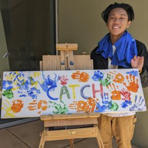 Teenage boy with medium skin tone and dark hair smiles as he poses next to white canvas painted with word "CATCH" and brightly colored hand prints