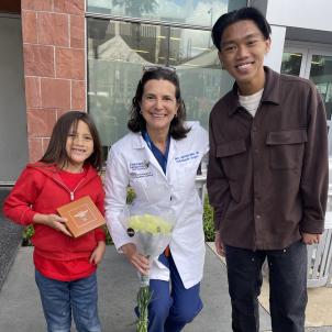 A young boy with medium skin tone and long brown hair wearing a red sweatshirt smiles he poses with a female doctor with light skin tone and dark hair wearing a white doctor's coat and a teenage boy with medium skin tone and dark hair wearing a brown shirt