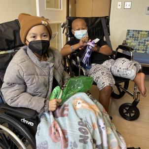 Two medium-skinned boys sit side by side in wheelchairs, smiling behind face masks, in a hospital waiting room.