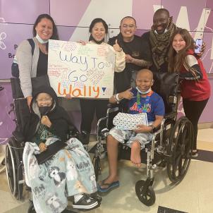 A group of four adults and a teenager stand smiling behind two boys in wheelchairs. Two of the women hold a sign, “Way to Go Wally!”
