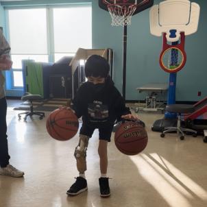 A dark-haired boy in a sweatshirt and shorts bounces two basketballs at the same time in a gym. His right leg has a prosthesis.