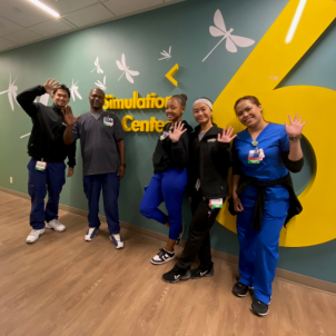 Five casually dressed nurse trainees smile and wave as the stand against CHLA's Simulation Center signage in a hospital corridor