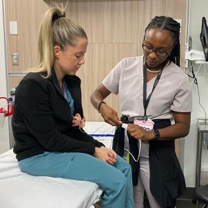A nurse with light skin tone and blonde hair sits on a hospital bed as a nurse with dark skin tone and dark hair adjusts cardiac leads