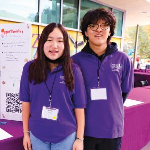 Teenage boy with medium skin tone and dark hair wearing a purple shirt and long black pants, standing next to a light skin tone teenage girl wearing a purple shirt and jeans at an outdoor event.