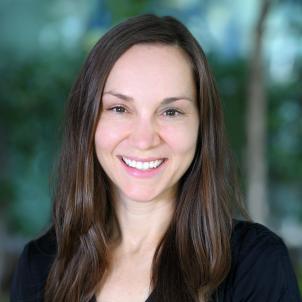 A woman with light skin tone and long dark hair smiles against a blurred outdoor background