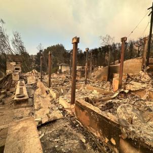 Charred concrete rubble, ashes, and metal beams stand next to burned trees and powerlines under a smoke-filled sky.
