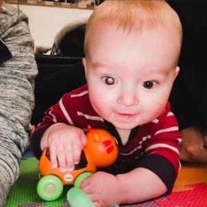 A baby with a light skin tone and a discolored right pupil, holding an orange toy.
