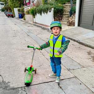 A young boy wearing a helmet stands next to his scooter in an alley 
