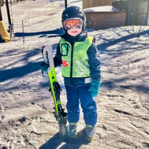 A young boy stands in front of ski slopes holding skis. 