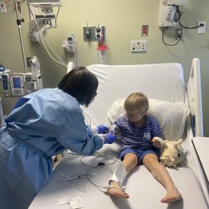 A toddler sits on a hospital bed while an infusion nurse places his infusion.