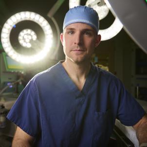 A man with light skin tone wearing dark blue scrubs and a light blue surgical cap gazes at the camera with a serious expression in an operating room. Two circular white bright lights are behind him.