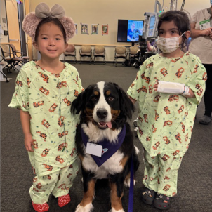 Two young patients wearing matching hospital scrubs pose with a dog therapy volunteer, a Bernese Mountain Dog, at Dog Therapy Appreciation Day.
