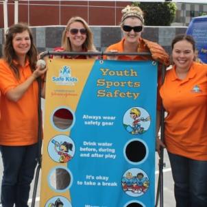 Four women stand smiling behind a banner from Safe Kids Los Angeles with information about youth sports safety.