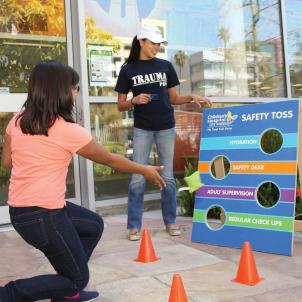 A medium skin-toned young woman tosses a bean bag at a board labeled Safety Toss, with different choices next to each hole in the board. 