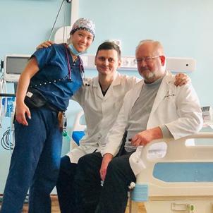 A light-medium skin-toned woman in blue scrubs with a surgical cap poses with two light skinned men wearing lab coats