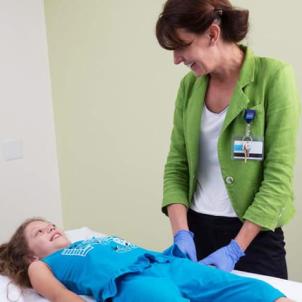 A light skin-toned young girl laying on an exam table and a light skin-toned woman wearing a green jacket and ID badge smile at each other.