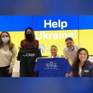 A light skin-toned woman in a surgical mask, a medium skin-toned woman in a surgical mask holding a T-shirt with COWF on it, a medium skin-toned woman holding a T-shirt with a handprint logo on it, a light skin-toned man and a medium-light skin-toned woman pose smiling in front of a sign that say Help Ukraine!