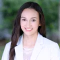 Headshot of woman with light skin tone and dark hair wearing white suit jacket with blurred outdoor background