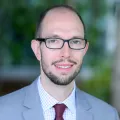 Headshot of a smiling man with light skin tone and receding dark hair wearing glasses and a purple tie and light blue suit jacket against a blurred outdoor background