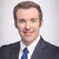 Headshot of a smiling man with light skin tone and brown hair wearing a blue tie and grey suit jacket against a neutral background