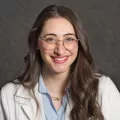 Headshot of a smiling woman with light skin tone and dark hair wearing white lab coat