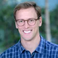 Headshot of a smiling man with light skin tone and light brown hair wearing tortoise shell glasses and a blue plaid shirt against a blurred outdoor background