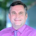 Headshot of a smiling man with light skin tone and short grey hair wearing a fuchsia dress shirt and purple tie against a blurred outdoor background