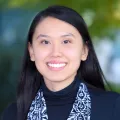 Headshot of a smiling young woman with medium skin tone and long dark hair wearing a dark blue turtleneck and patterned scarf against a blurred background