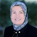 Headshot of a smiling woman with light skin tone wearing a light blue patterned headscarf and dark jacket against a blurred outdoor background