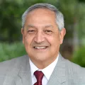 Headshot of a smiling man with medium skin tone and grey hair wearing a white dress shirt and maroon tie under a grey suit jacket against a blurred outdoor background