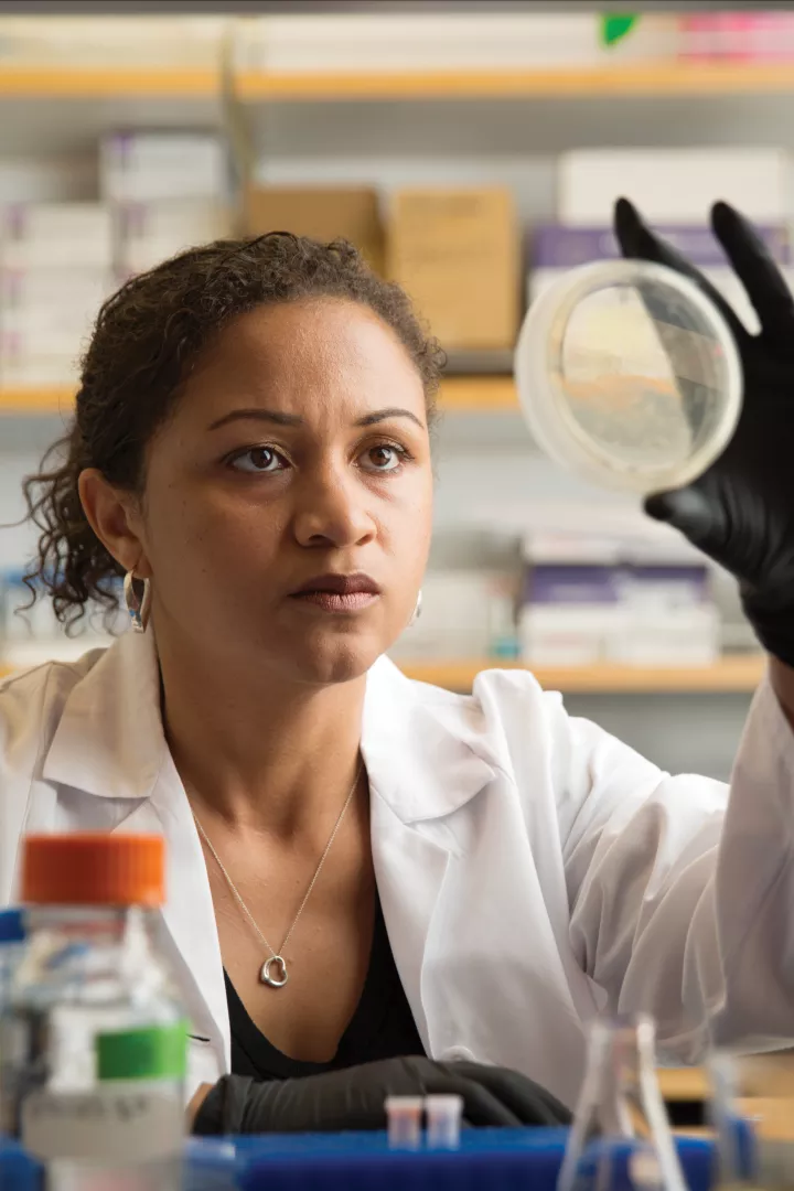 A woman with medium-dark skin tone wearing a lab coat holds a dish up to look at a research sample.