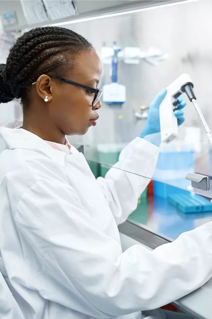 A woman with dark skin tone and braids tucked into a bun wears a lab coat and uses a pipette device in a laboratory fume hood.