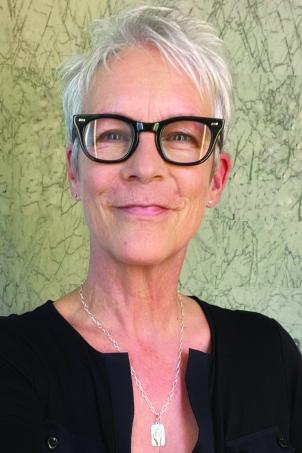 Actress Jamie Lee Curtis, a woman with light skin tone and frosty hair, wearing black-framed glasses smiles while molding a silver necklace