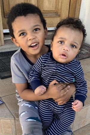 Young boy with medium skin tone and dark hair wearing pajamas sits on stoop while holding toddler with medium skin tone and dark hair wearing a onesie