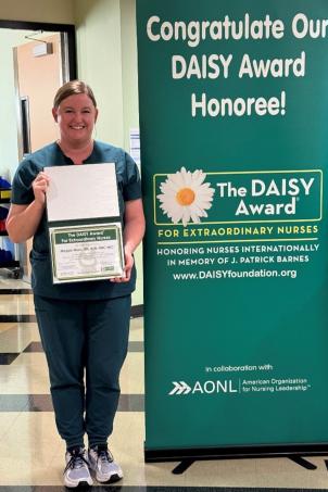 Female nurse with light skin tone and blonde hair smiles as she poses with their DAISY Award next to a DAISY Award poster