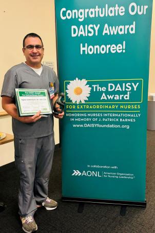 Male nurse with medium skin tone and short dark hair smiles as he poses with his DAISY Award next to a DAISY Award poster