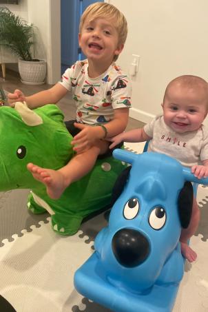 A light skin tone male toddler with blonde hair sitting on a green bouncy dinosaur toy playing with a light skin tone baby sitting on a blue bouncy dog toy