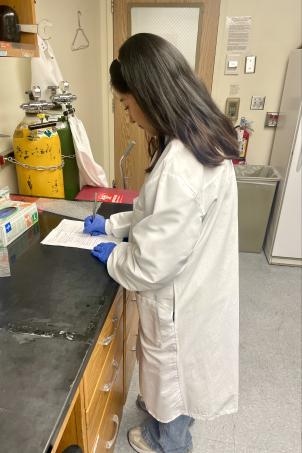 A young adult with a medium skin tone wears a white lab coat and gloves and stands at a black table in a laboratory, writing on a piece of paper.