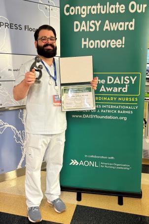 Male nurse with medium skin tone, dark hair, full beard and glasses smiles as they pose with their DAISY Award next to a DAISY Award poster