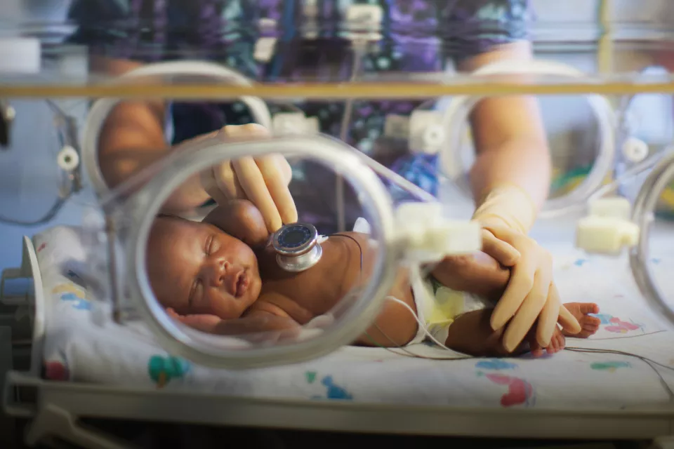 A woman with light skin tone holds a stethoscope on the chest of a medium-dark skin toned infant in an incubator.