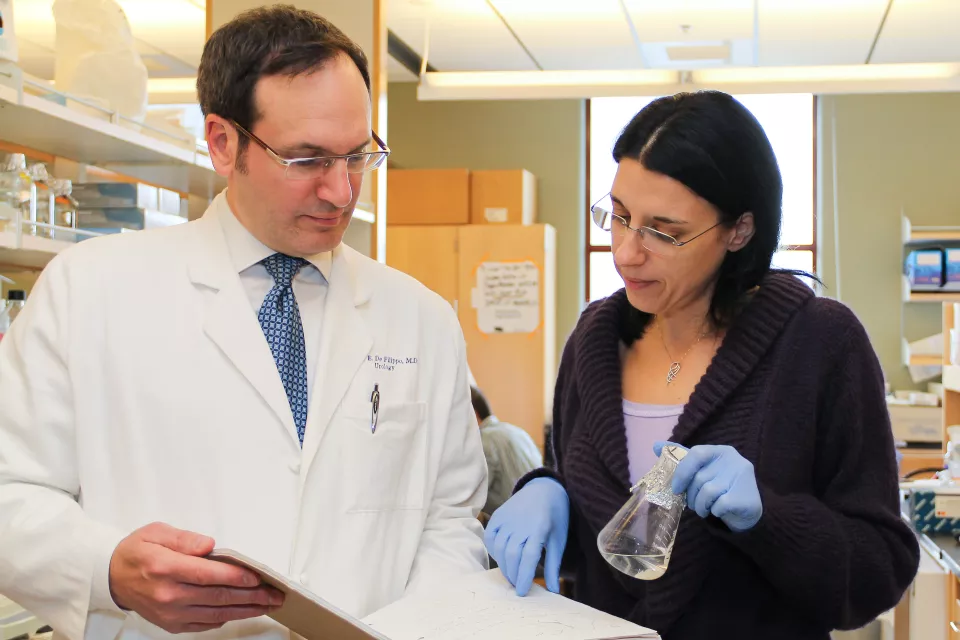 A woman with light skin tone holds a glass beaker covered with foil. She points into a notebook held open by a light skin toned man in a lab coat.