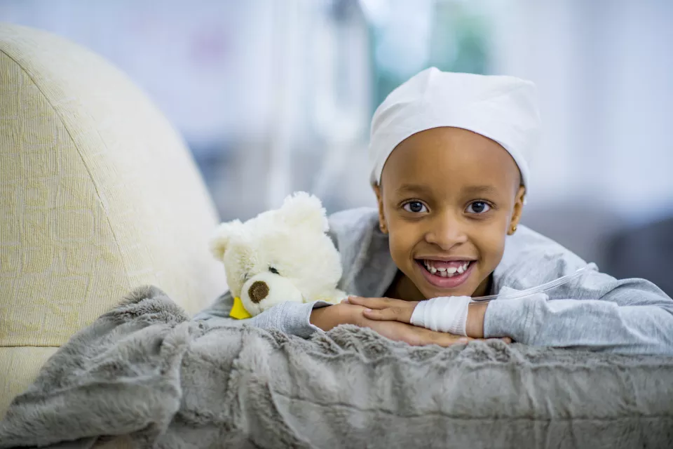 A little girl with medium skin tone lays on a cushion, smiling. She holds a teddy bear and wears a headscarf. A tube is bandaged to the back of her hand.