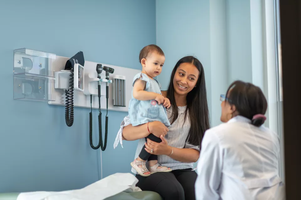 A young woman with medium skin tone sits on exam table holding a toddler. A doctor with medium-dark skin tone is speaking to them.